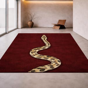 Op de afbeelding: Een rechthoekig, dieprood tapijt met een groot, gestileerd slangenontwerp. De slang heeft bruine, zwarte en beige markeringen, die contrasteren met de effen achtergrond. Het tapijt ligt in een moderne, minimalistische kamer.
