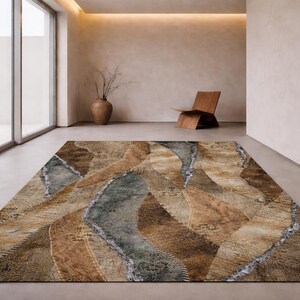 May include: A rectangular area rug with a unique, abstract design. The rug features a blend of brown, tan, and gray tones, with a wavy pattern and stitched details. The rug is placed on a light-colored floor in a modern room.
