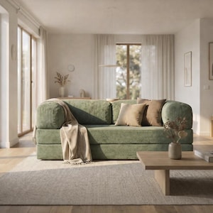 Può includere: Un divano in velluto a coste verde salvia con braccioli arrotondati e diversi cuscini beige e marroni. Una coperta beige è drappeggiata sul braccio. Il divano è su un tappeto grigio chiaro in un soggiorno luminoso.