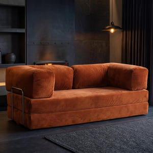 May include: A contemporary sofa in a warm, rust-orange corduroy fabric. The sofa has a low, boxy design with square armrests and a metal frame detail. It's set in a modern living room with a dark grey rug in front. The sofa measures approximately 200 cm in length.