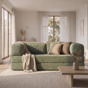 May include: A sage green corduroy sofa with rounded arms and back. The sofa has several brown throw pillows and a beige blanket draped over the arm. A wooden coffee table and a vase of dried flowers are in front of the sofa.