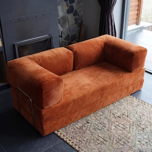May include: A burnt orange corduroy sofa with a boxy design. The sofa has two armrests and a low profile. The frame is visible on the sides. The sofa sits on a patterned rug. A fireplace and window are in the background.