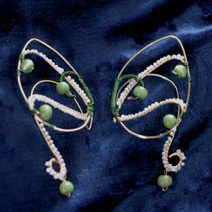 May include: A pair of handcrafted ear cuffs with a whimsical, elven design. The ear cuffs feature a gold-colored wire frame, adorned with green beads and delicate white beading. Green wire wraps add detail. The design is set against a dark blue background.