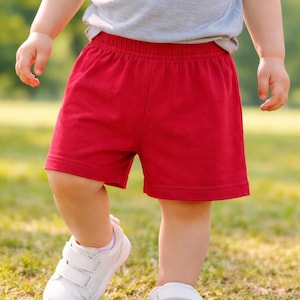 May include: A toddler wearing a gray t-shirt, red shorts, and white sneakers with velcro straps is captured in a medium shot. The child is walking on a grassy surface, with a blurred green background.