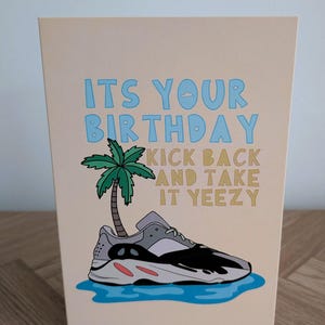 May include: A birthday card with the text "ITS YOUR BIRTHDAY KICK BACK AND TAKE IT YEEZY" in blue and gold. The card features a cartoon sneaker, a palm tree, and a blue water design on a beige background.