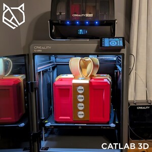 May include: A red and gold 3D-printed gift box with a gold bow, inside a Creality K2 Plus 3D printer. The printer has a control panel and a Creality CFS unit on top. The text "CATLAB 3D" is at the bottom.