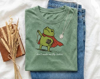 May include: Sage green t-shirt featuring a cartoon frog in a cape, holding a rosary and book. The text "don't make me flip tables" is printed below. The shirt is styled with blue jeans and a silver pendant necklace.