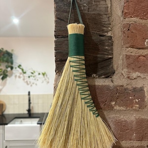 Può includere: Una scopa fatta a mano con una testa di paglia naturale e un manico verde avvolto. Il manico è fissato con cuciture verdi. La scopa è appesa a una trave di legno contro un muro di mattoni. Un oggetto decorativo.