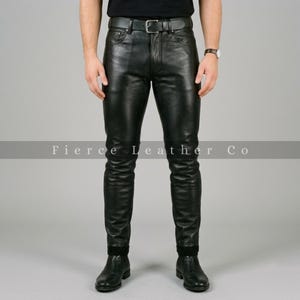 May include: Black leather trousers featuring a classic five-pocket design, a front button and zip fastening, and a matching black leather belt with a silver-toned metal buckle. The trousers are worn with black leather boots and a black shirt.