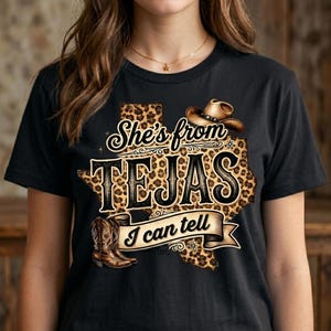 May include: Black t-shirt with a graphic featuring a Texas-shaped design with a leopard print pattern. The text reads "She's from TEJAS I can tell" with a cowboy hat and boots illustration.