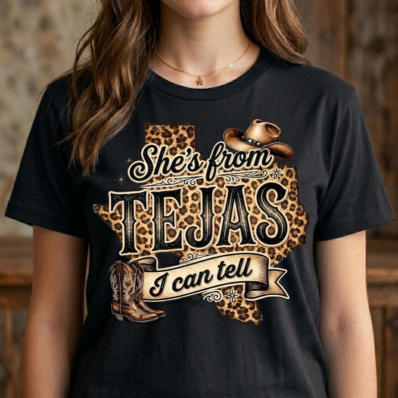 May include: Black t-shirt with a graphic featuring a Texas-shaped design with a leopard print pattern. The text reads "She's from TEJAS I can tell" with a cowboy hat and boots illustration.