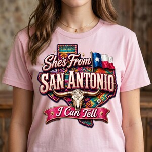 May include: Pink t-shirt featuring a graphic design with the text "She's From San Antonio I Can Tell". The design includes a Texas outline, a steer skull, and the Texas state flag.
