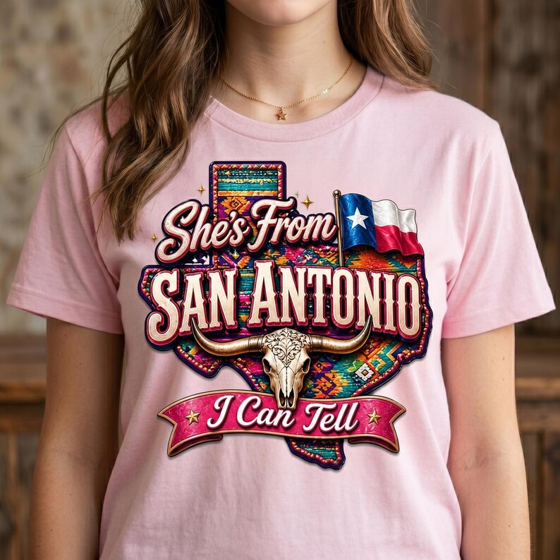 May include: Pink t-shirt featuring a graphic design with the text "She's From San Antonio I Can Tell". The design includes a Texas outline, a steer skull, and the Texas state flag.