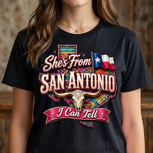 May include: Black t-shirt with a colorful graphic design. The design features the text "She's From San Antonio" with a Texas-shaped outline, a steer skull, and a Texas flag. The phrase "I Can Tell" is also included.
