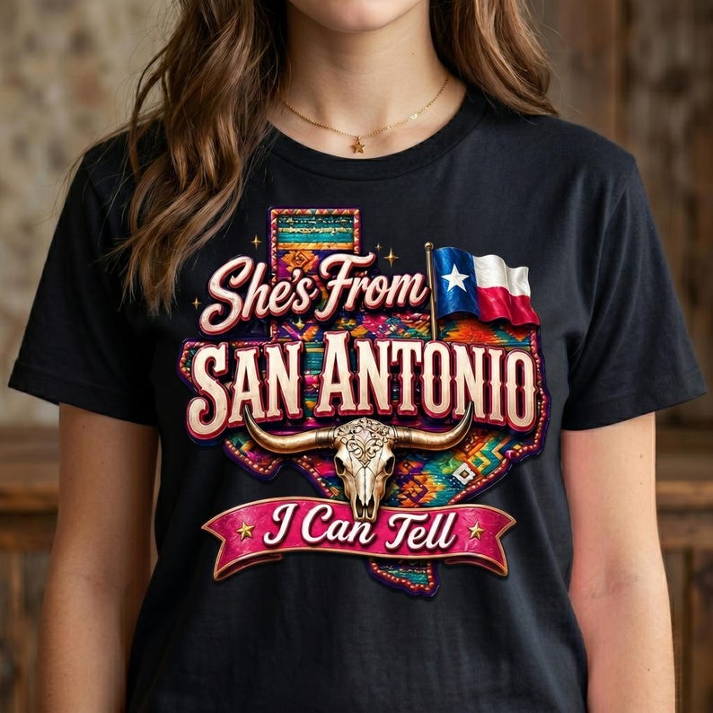 May include: Black t-shirt with a colorful graphic design. The design features the text "She's From San Antonio" with a Texas-shaped outline, a steer skull, and a Texas flag. The phrase "I Can Tell" is also included.