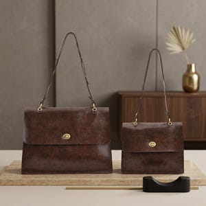 May include: Two dark brown leather handbags of different sizes, each featuring a rectangular flap, a gold-toned turn-lock closure, and a thin shoulder strap attached with gold-toned hardware. The bags have a smooth, slightly distressed finish.