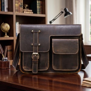 Könnte beinhalten: Braune Leder-Umhängetasche mit zwei Vordertaschen und einem Klappenverschluss. Die Tasche hat einen Schultergurt und einen Schnallenverschluss. Das Leder hat einen Used-Look mit sichtbaren Nähten. Die Tasche steht auf einer Holzoberfläche.