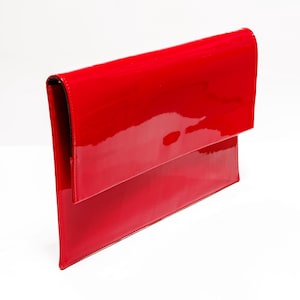 Può includere: Pochette rossa lucida con patta. La borsa ha una forma rettangolare e una finitura lucida, simile alla vernice. La pochette è su sfondo bianco.