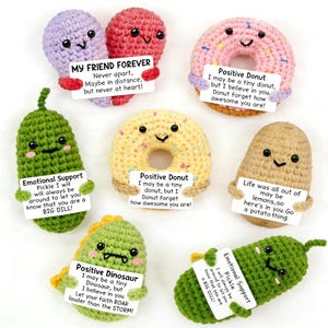 Op de afbeelding: Een verzameling handgemaakte gehaakte knuffels in verschillende vormen, waaronder een augurk, een dinosaurus, een aardappel en donuts. Elke knuffel houdt een klein bordje vast met positieve boodschappen. Het kleurenpalet omvat groen, roze, geel en paars.