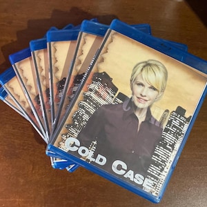 May include: A stack of blue Blu-ray cases featuring the television series Cold Case. The cover art displays a blonde person in a dark shirt against a city skyline background with a tan, parchment-like border. The title Cold Case is printed in white.