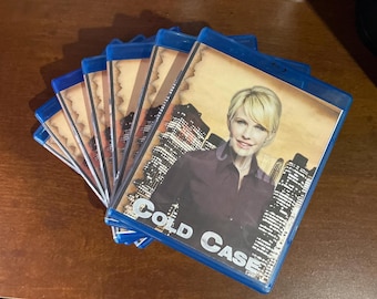 Könnte beinhalten: Ein Stapel blauer Blu-ray-Hüllen der Fernsehserie Cold Case. Das Cover zeigt eine blonde Person in einem dunklen Hemd vor einer nächtlichen Skyline mit einem beigen, pergamentartigen Rand. Der Titel Cold Case ist in weißer Schrift gedruckt.