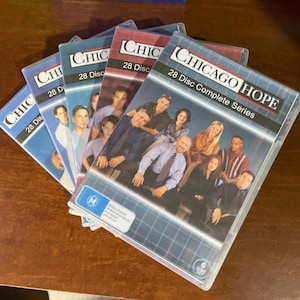Puede incluir: Un conjunto de estuches de DVD de la serie de televisión "Chicago Hope". Los estuches son de plástico transparente con diseños azules, rojos y blancos. La parte frontal de los estuches muestra imágenes del elenco y el texto "28 Disc Complete Series".