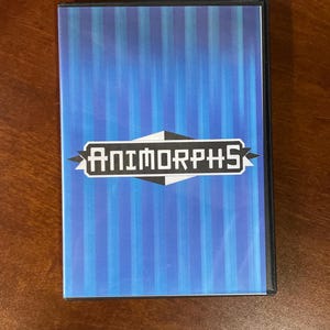 Puede incluir: Una caja de DVD con un fondo a rayas azules y blancas. El título "Animorphs" se muestra en blanco, con una fuente estilizada, dentro de una forma geométrica en blanco y negro. La caja es negra.