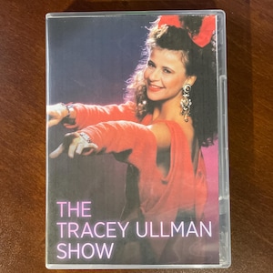 May include: A VHS case for "The Tracey Ullman Show" with a woman in a red dress and a red bow. The show's title is in white and pink text on a dark background.