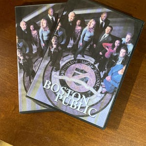 Könnte beinhalten: Zwei DVD-Hüllen mit der Fernsehserie "Boston Public". Die Hüllen zeigen eine Gruppe von Personen, mit dem Titel der Serie und dem Text "Winslow High School". Die Hüllen liegen auf einer Holzoberfläche.