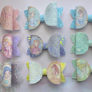 May include: A collection of nine hair bows featuring various cartoon characters in circular frames. Each bow is crafted from glittery material with a lace overlay in pastel colours including pink, blue, purple, green, and yellow.