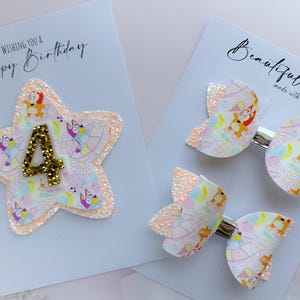 May include: A star-shaped hair accessory with a gold glitter number 4 and two matching hair bows, all featuring a light-coloured pattern of cartoon dogs and flowers on a white background. The items are displayed on white cards with text.