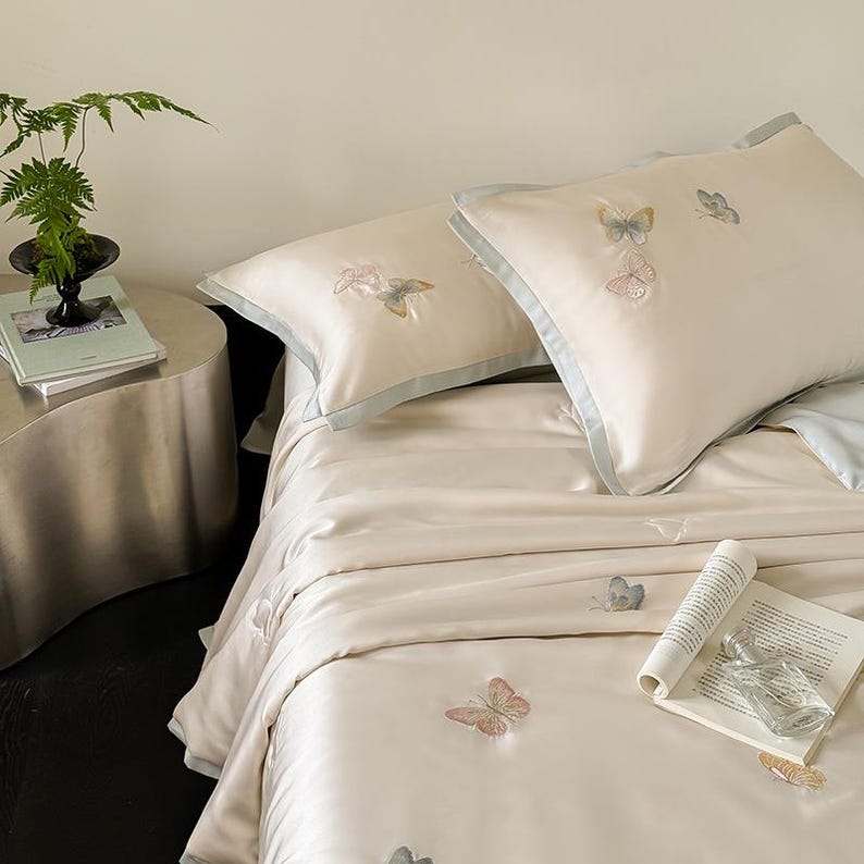 Puede incluir: Juego de cama color crema con mariposas bordadas. El juego incluye dos almohadas con ribete azul claro y una funda n&oacute;rdica a juego. Un libro y un peque&ntilde;o jarr&oacute;n con vegetaci&oacute;n est&aacute;n sobre una mesita auxiliar.