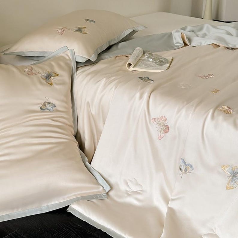 Puede incluir: Juego de cama de sat&eacute;n color crema con bordados de mariposas rosas, azules y doradas. El juego incluye fundas de almohada y una funda n&oacute;rdica. Una revista enrollada descansa sobre la cama. La ropa de cama tiene un ribete azul claro.