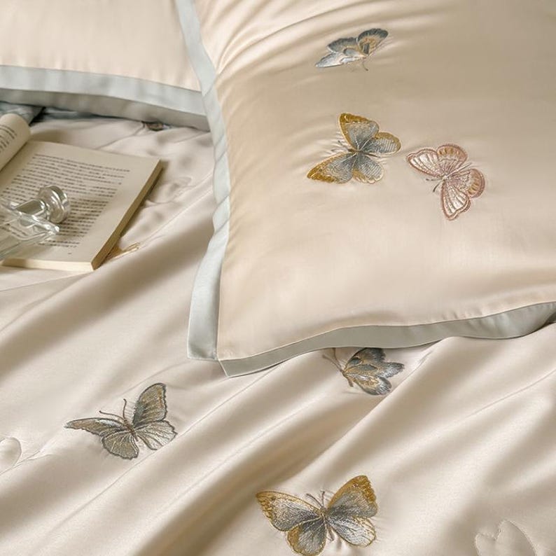 Puede incluir: Juego de cama color crema con detalles de mariposas bordadas. El juego incluye una funda de almohada con borde azul claro y varias mariposas bordadas en tonos dorados, grises y rosas. Tambi&eacute;n se ven un libro y un objeto decorativo.