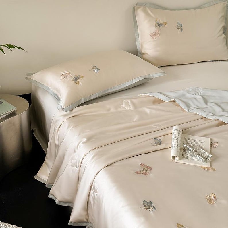 Puede incluir: Juego de cama beige con detalles de mariposas bordadas. El juego incluye una funda n&oacute;rdica, dos fundas de almohada y una s&aacute;bana bajera. Las fundas de almohada y la funda n&oacute;rdica tienen un ribete azul claro. Una revista enrollada y un frasco de perfume est&aacute;n sobre la cama.