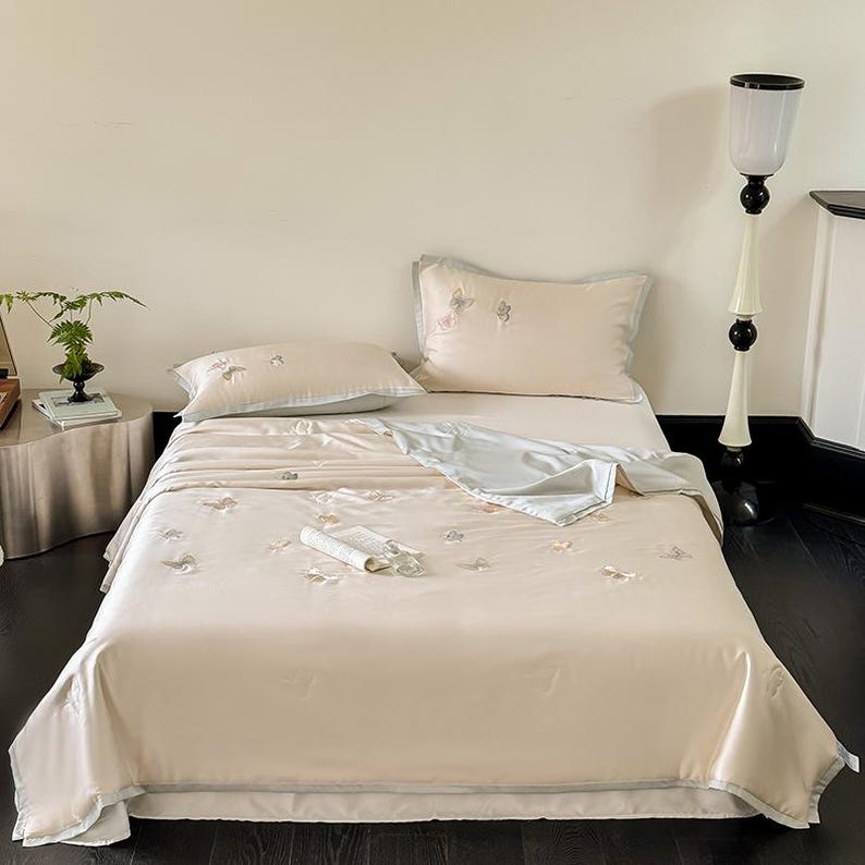 Puede incluir: Un juego de cama beige con detalles de mariposas bordadas. El juego incluye una s&aacute;bana bajera, una s&aacute;bana encimera y dos fundas de almohada. Una manta azul claro est&aacute; doblada encima. La cama est&aacute; hecha y lista para usar.