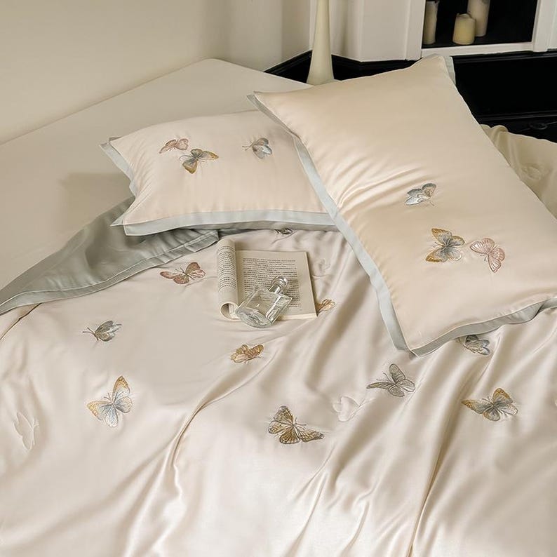 Puede incluir: Juego de cama color crema con detalles de mariposas bordadas. El juego incluye dos almohadas con ribete azul claro, una funda n&oacute;rdica y una s&aacute;bana bajera. Un libro y un frasco de perfume est&aacute;n sobre la cama, agregando elegancia.