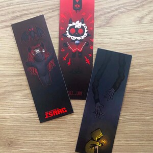 May include: Three rectangular bookmarks featuring video game illustrations. One shows a character with red hair and the text THE BINDING OF ISAAC. Another depicts a white lamb character with the text CULT OF THE LAMB. The third shows a figure in a yellow hood.