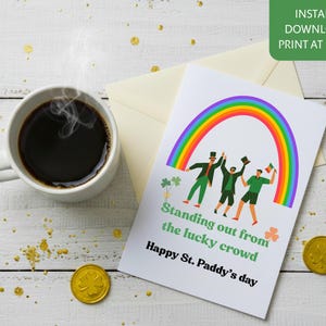 Może przedstawiać: Kartka z okazji Dnia Świętego Patryka z tęczą, skrzatami i tekstem "Standing out from the lucky crowd". Filiżanka kawy, złote monety i koniczyny na białej drewnianej powierzchni.