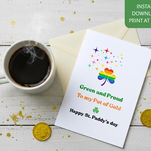 Może przedstawiać: Kartka z okazji Dnia Świętego Patryka z tęczową koniczyną i napisem "Green and Proud to my Pot of Gold". Na białej drewnianej powierzchni filiżanka kawy, złote monety i zielona koniczyna.