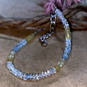 May include: A delicate bracelet featuring small, round beads in shades of clear, pale blue, and yellow. The bracelet has a silver-colored chain and clasp. The beads are strung together to form a flexible band, perfect for everyday wear.