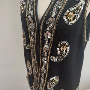 May include: A black sleeveless waistcoat featuring intricate embroidery and embellishments in silver and gold sequins. The garment displays a repeating paisley pattern along the front opening and scattered across the body, with a V-neckline design.