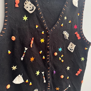 Op de afbeelding: Een zwart gebreid vest met een V-hals en een knoopsluiting aan de voorzijde. Het kledingstuk is versierd met diverse Halloween-thema applicaties, waaronder stoffen spookjes, pompoentasjes, oranje snoepjes en kleurrijke geborduurde sterren.