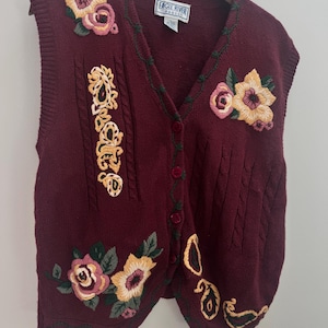 Può includere: Un gilet in maglia bordeaux senza maniche con scollo a V e chiusura frontale a bottoni. Il capo è decorato con motivi floreali ricamati in tonalità di giallo, rosa e verde, insieme a dettagli a trecce. Un'etichetta riporta la scritta Eagle River.