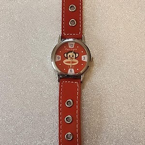 Może przedstawiać: Czerwony zegarek na rękę ze srebrną kopertą i czerwonym skórzanym paskiem. Tarcza zegarka jest czerwona z napisem "Paul Frank" i grafiką małpy z kreskówek. Pasek ma srebrne nity.