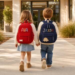May include: Two corduroy backpacks, one red with the name AVA and a small bow, and one navy blue with the name JAXON and a dinosaur patch. Both bags feature front zippered pockets, top handles, and adjustable shoulder straps for carrying.