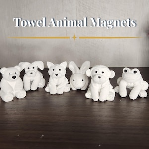 May include: A collection of white towel animal magnets, including a bear, two dogs, a rabbit, a monkey, and a frog. Each animal has black dot eyes and is arranged on a dark brown surface. The text "Towel Animal Magnets" is at the top.