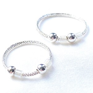 May include: Two circular rings featuring a textured silver-toned band. Each ring is accented with two polished silver-toned spherical beads and two small, translucent white cube-shaped beads arranged in a repeating pattern along the band.