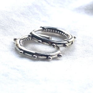 Könnte beinhalten: Ein Paar silberne Creolen mit polierter Metalloberfläche und einer Reihe kleiner, gleichmäßig verteilter kugelförmiger Perlen entlang der Außenkante jedes kreisförmigen Rings. Die Creolen verfügen über einen Klappverschluss und eine texturierte Innenseite.