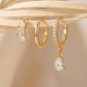May include: Three pairs of gold earrings. One pair has a cluster of marquise-cut cubic zirconia stones. Another pair is a simple hoop design. The third pair has a teardrop-shaped cubic zirconia pendant.